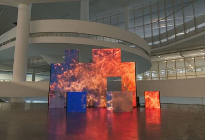 Imagem do interior de um museu com pé direito alto, vários vidros na paredes, uma rampa e várias telas digitais que formam uma imagem de nuvens e fogo. 