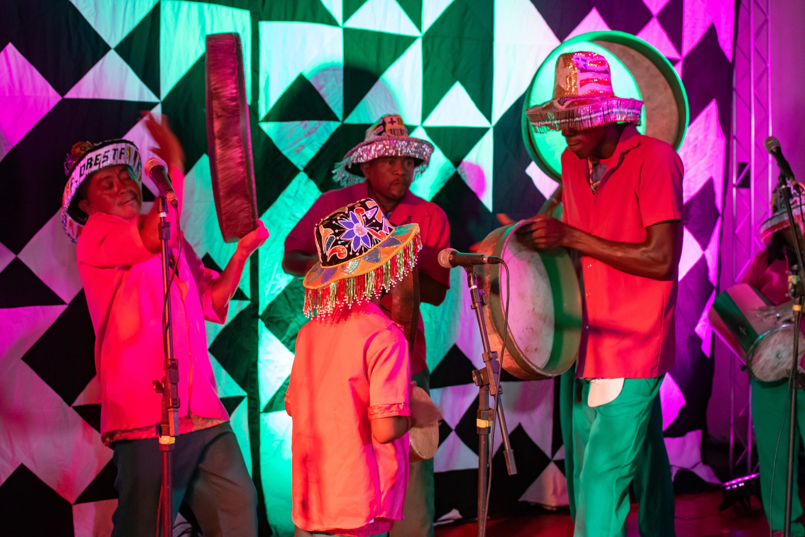 Foto de três homens e uma criança em cima de um palco com instrumentos musicais e microfone. Todos estão usando chapéu colorido.