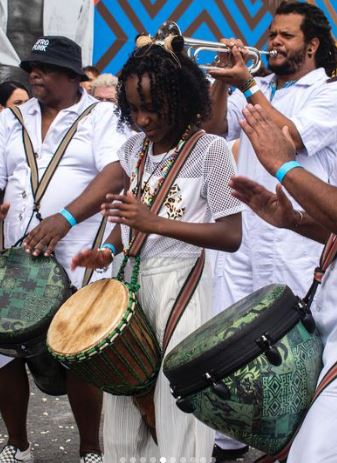 Foto de uma menina negra no centro da foto tocando um instrumento de percussão e outras pessoas em volta também e todos vestindo roupas brancas. 
