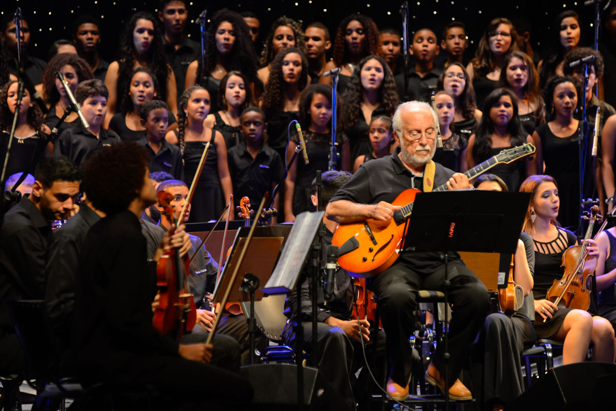 Foto de um homem tocando baixo, uma orquestra ao redor e crianças ao fundo cantando em um palco