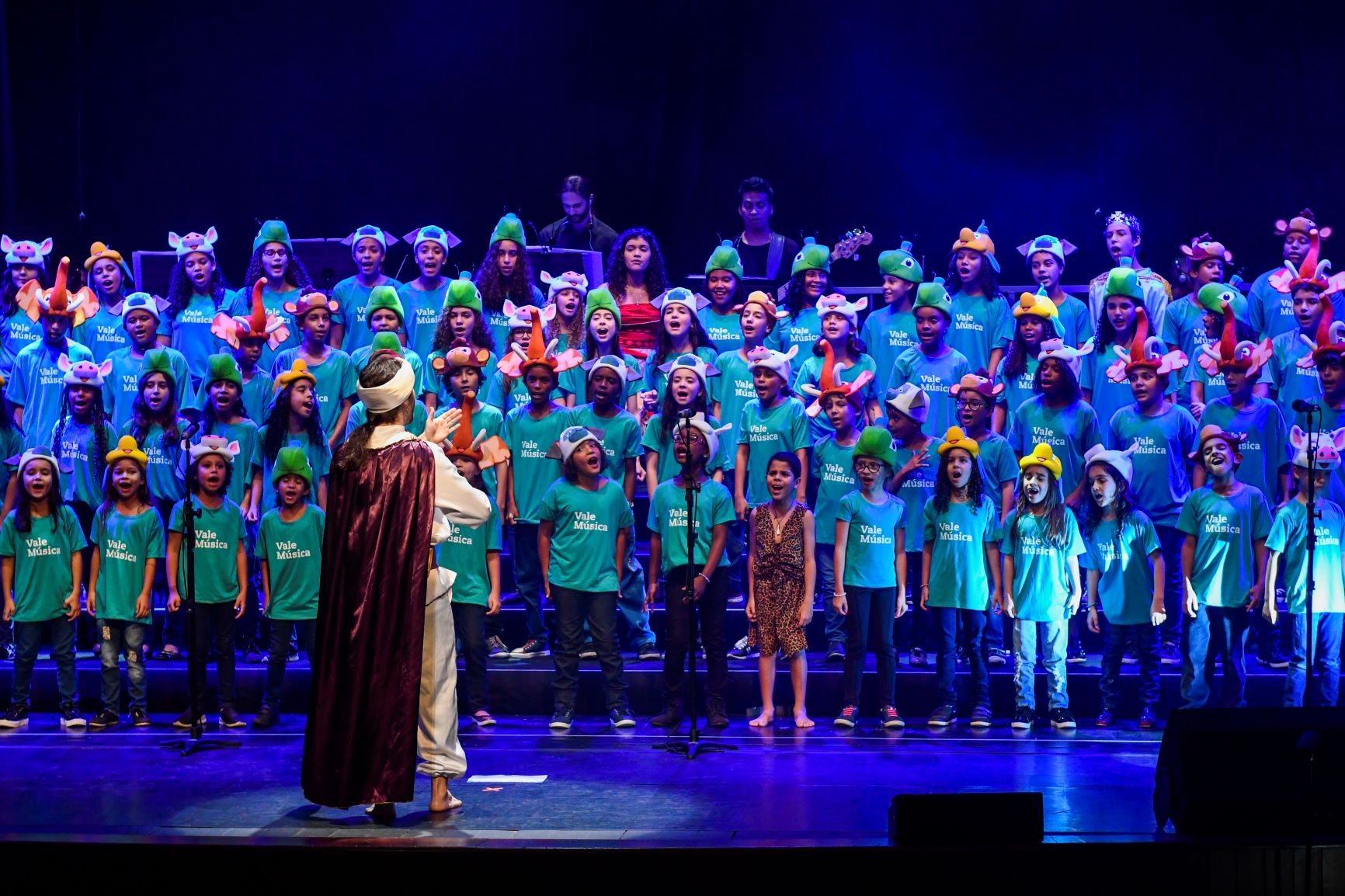 Foto de crianças no palco em uma apresentação com um professor como maestro na frente. Todas estão usando a camisa verde do Vale Música e estão com adereços na cabeça.