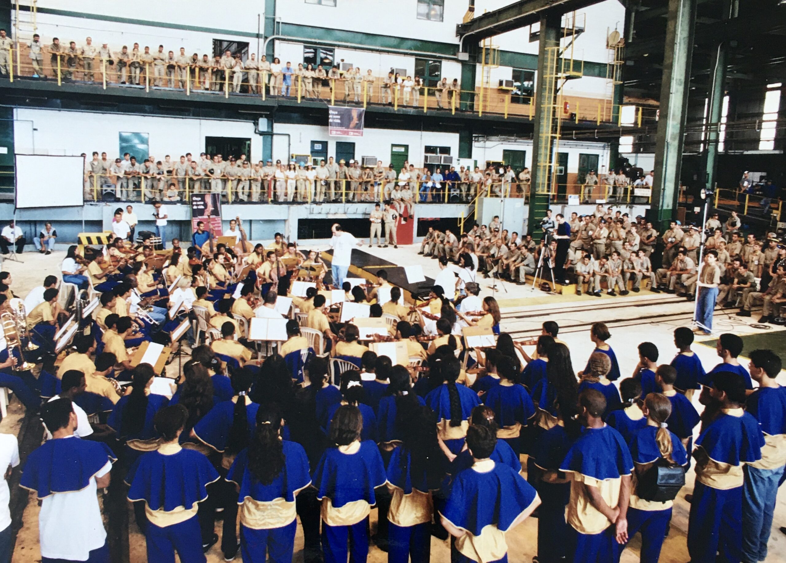 Foto de várias crianças e jovens cantando em um galpão e vários funcionários assistindo