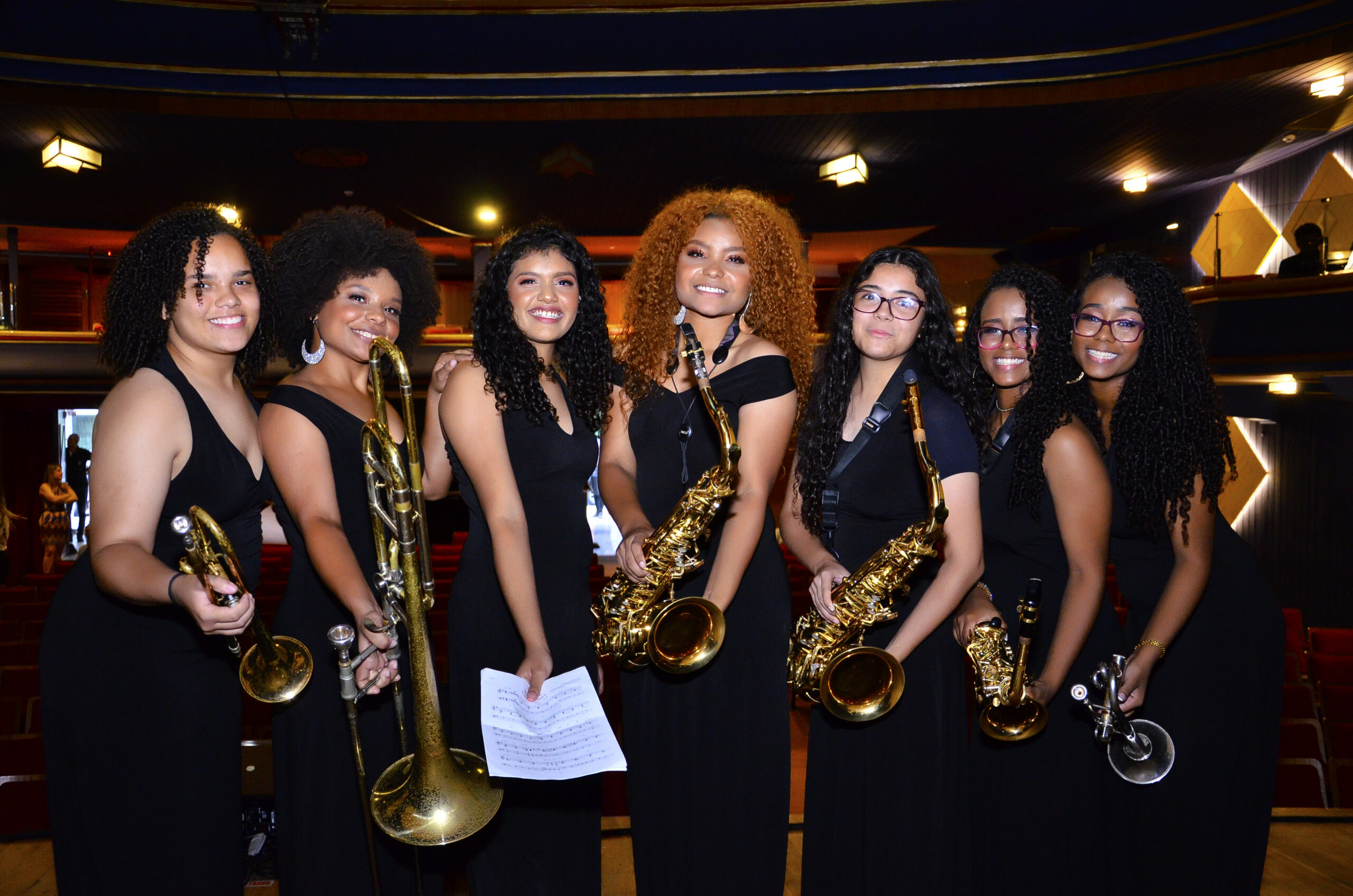 Foto de 7 mulheres negras com cabelos cacheados vestindo macacões pretos e cada uma segurando um saxofone