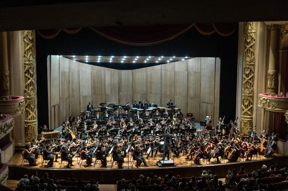 Foto de um palco de um teatro com uma orquestra sinfônica com vários músicos