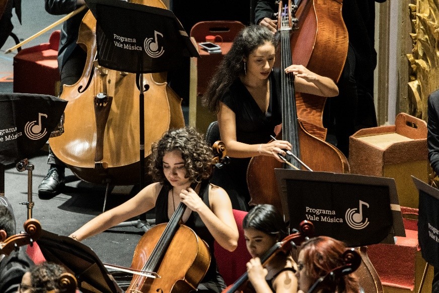 Foto com zoom de duas mulheres em um palco no meio de uma orquestra tocando violino