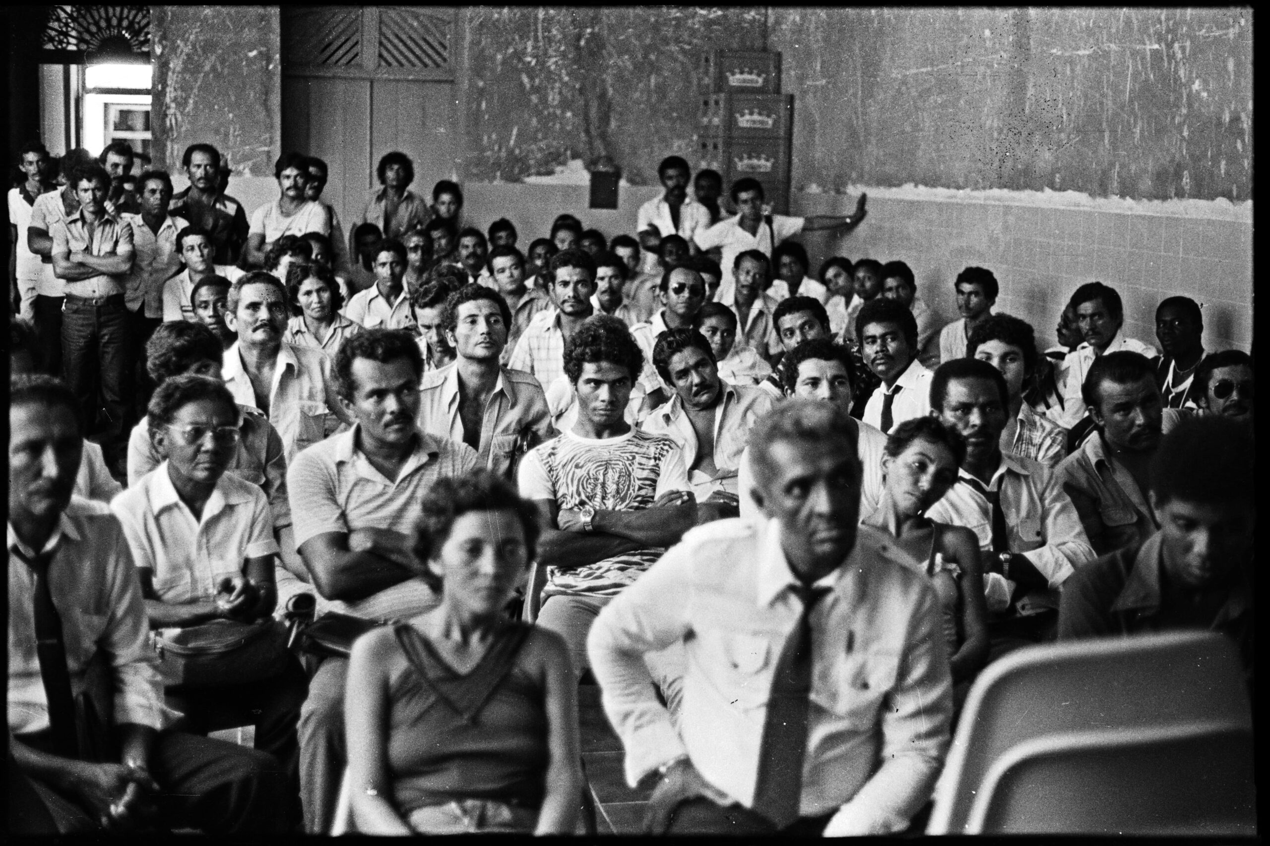 Foto preto e branca de diversas pessoas sentadas em uma sala, prestando atenção em algo que acontece na frente deles