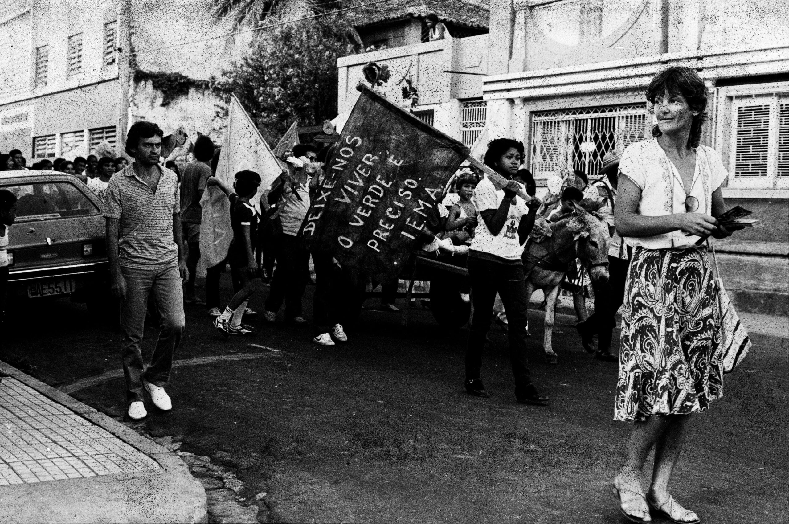 Imagem preto e branca de uma manifestação. Diversas pessoas caminham em uma rua segurando bandeiras. À frente está uma mulher.