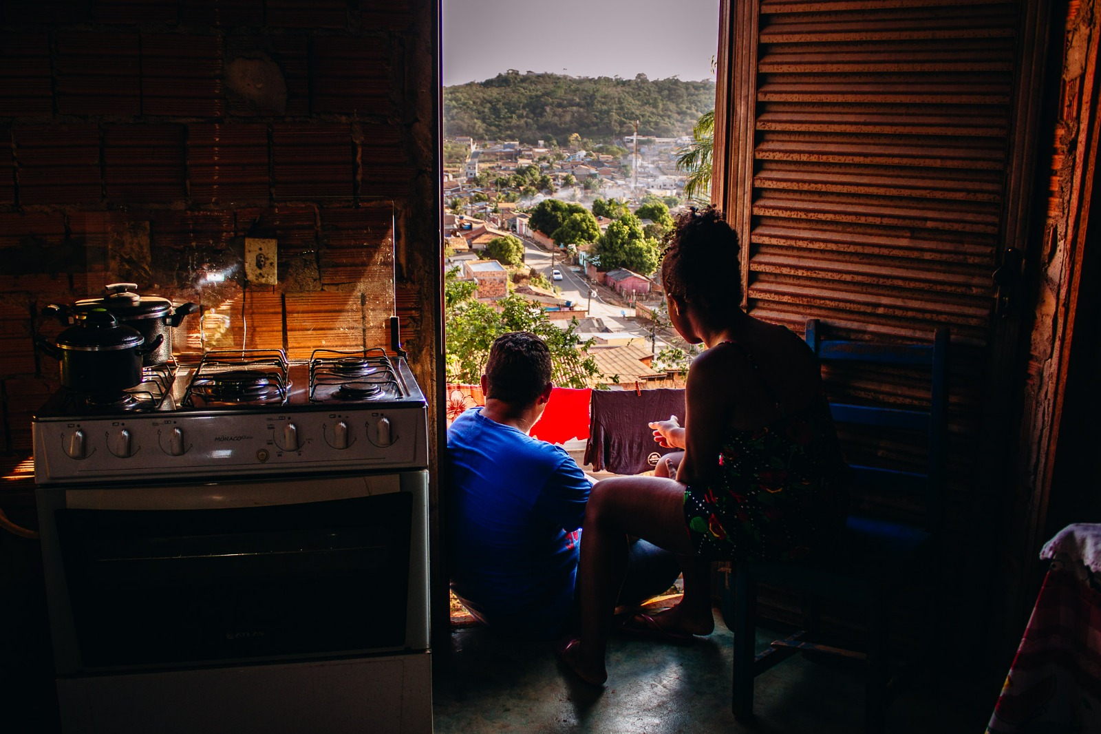 Uma mulher e um menino olham para o horizonte de casas através da porta de casa. No lado dentro é possível ver um fogão 