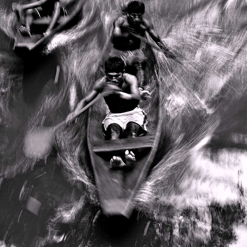 Dois homens sem camisa e com shorts sentados na canoa seguram remos com as mãos enquanto empurram a água para irem para a frente.