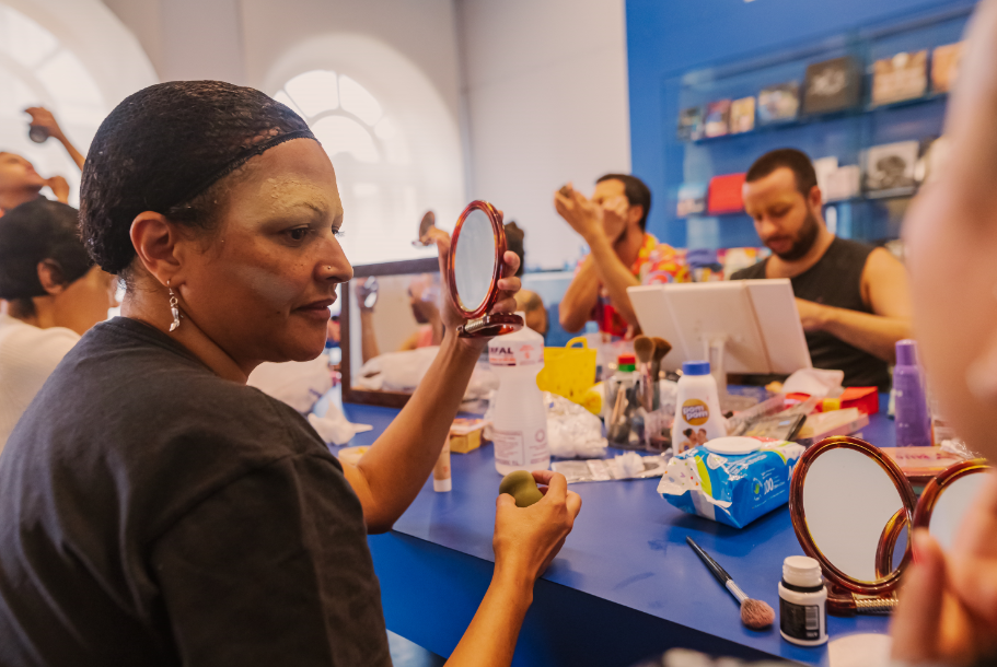 Pessoas sentadas no camarim seguram espelhos pequenos em frente aos seus rostos enquanto se maquiam para montação drag queen.