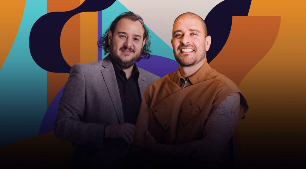 Dois homens de barba: um com cabelos na altura dos ombros com cachos nas pontas e outro sem cabelo com braços cruzados sorriem para a foto.