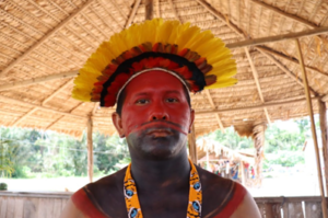 Homem indígena com o rosto e ombros pintados de vermelho e preto. Ele usa com cocar.